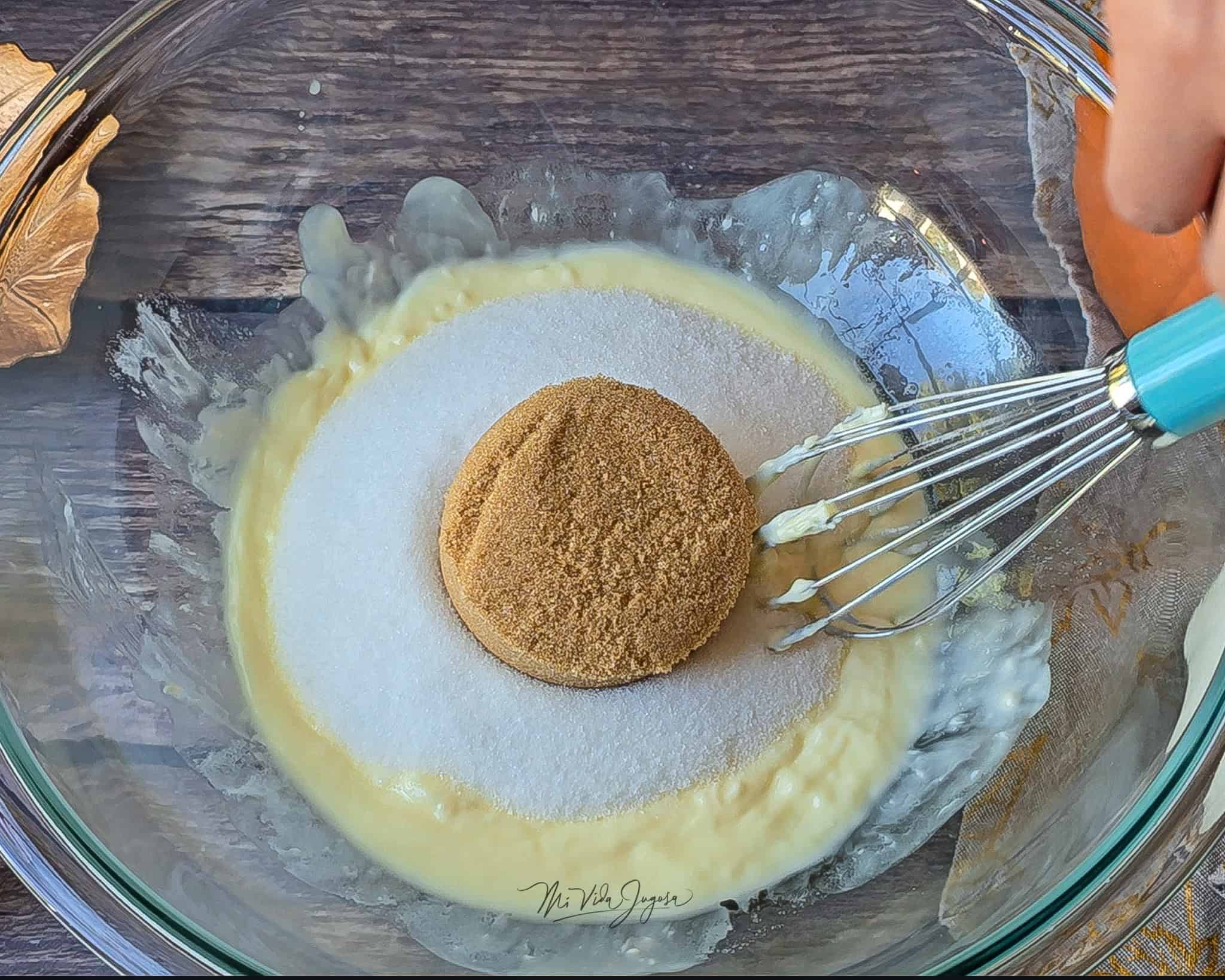 Tazón de vidrio con mantequilla derretida, azúcar blanca y azúcar morena por encima. Dentro del tazón se observa una espátula de globo, lista para mezclar los ingredientes.