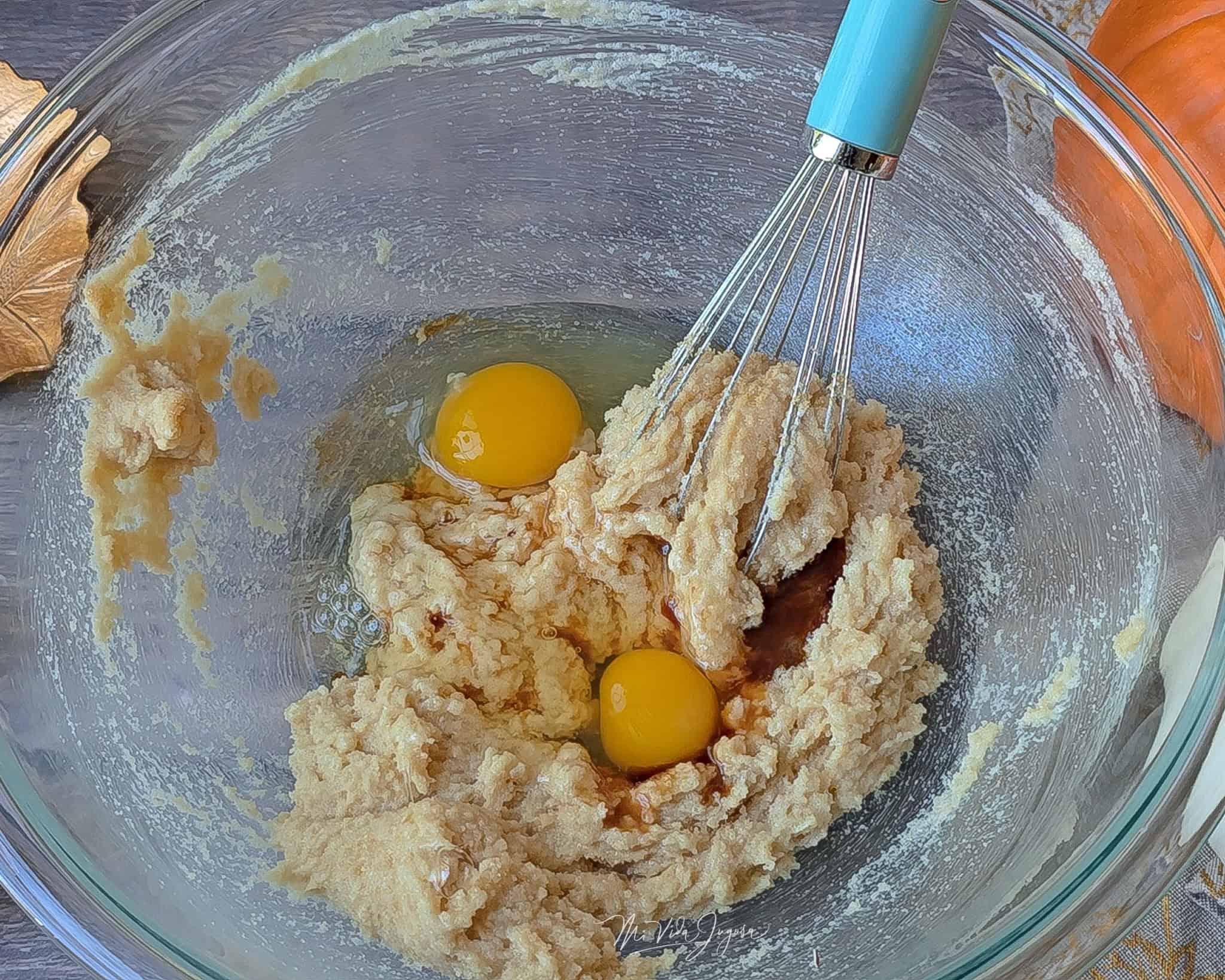 Tazón de vidrio con una mezcla de mantequilla y azúcar, con dos huevos encima y una espátula de globo dentro del tazón, listo para continuar con la preparación del queque de calabaza.
