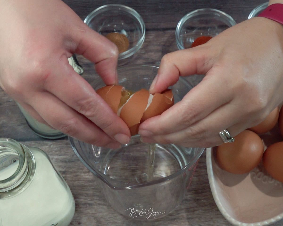 Unas manos abriendo la cáscara de un huevo sobre un envase de vidrio; al lado se observa una taza con huevos, otra con leche y unas tacitas con especias.