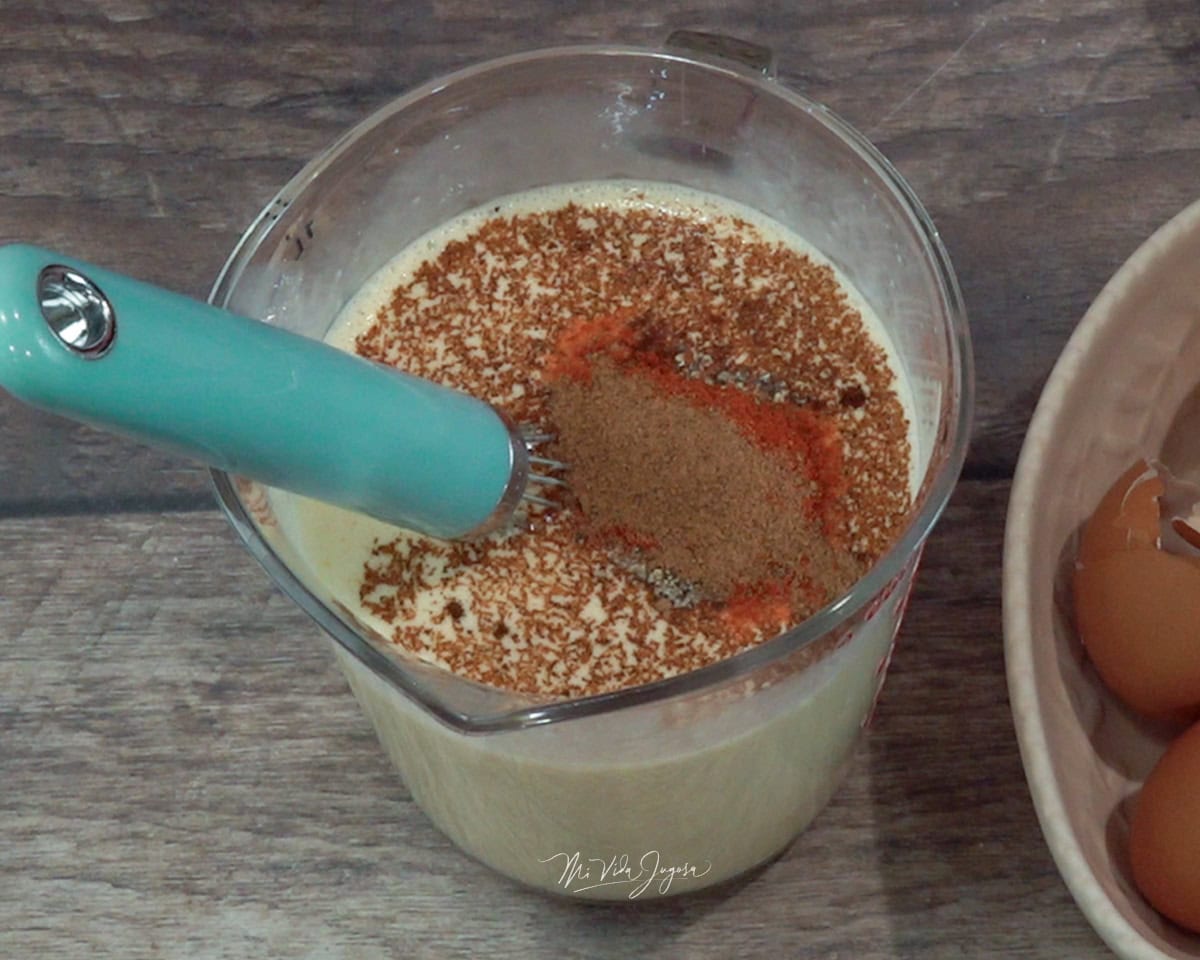 Una taza de medir con un batidor de globo dentro, conteniendo la mezcla de leche, paprika, nuez moscada y chile chipotle.