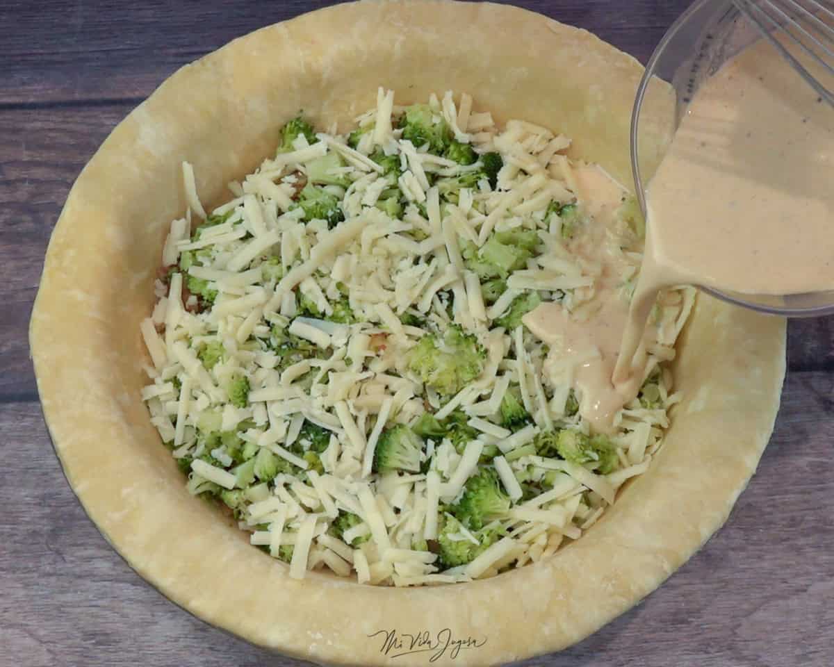 Un molde con una base de masa y relleno de brócoli con cebolla, mientras se agrega la mezcla líquida de huevo, leche, crema dulce y especias.