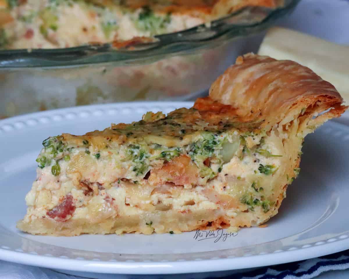 En la parte trasera de la imagen se visualiza un molde de quiche; en primer plano se observa un trozo de quiche con brócoli, tocineta y huevo.