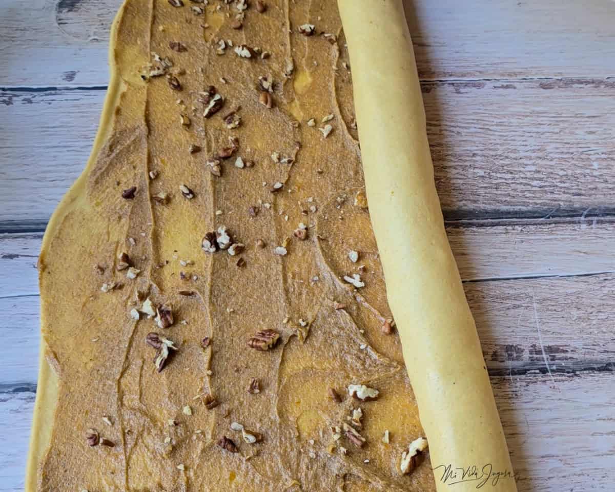 La masa estirada con una mezcla de azúcar y mantequilla por encima, mientras esta siendo enrollada desde un extremo.