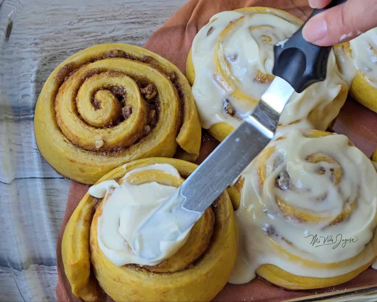Cuatro rollos de canela: dos cubiertos con glaseado de queso crema con sirope de maple, uno siendo cubierto con el glaseado usando una espátula plana y otro aún sin glasear.