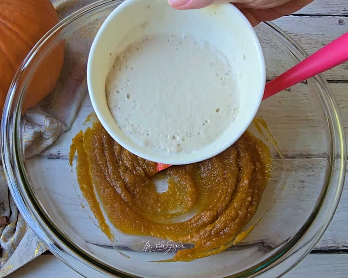 Un tazón con la mezcla de puré de calabaza y azúcar morena, mientras se agrega, desde una taza, la levadura activada en leche.