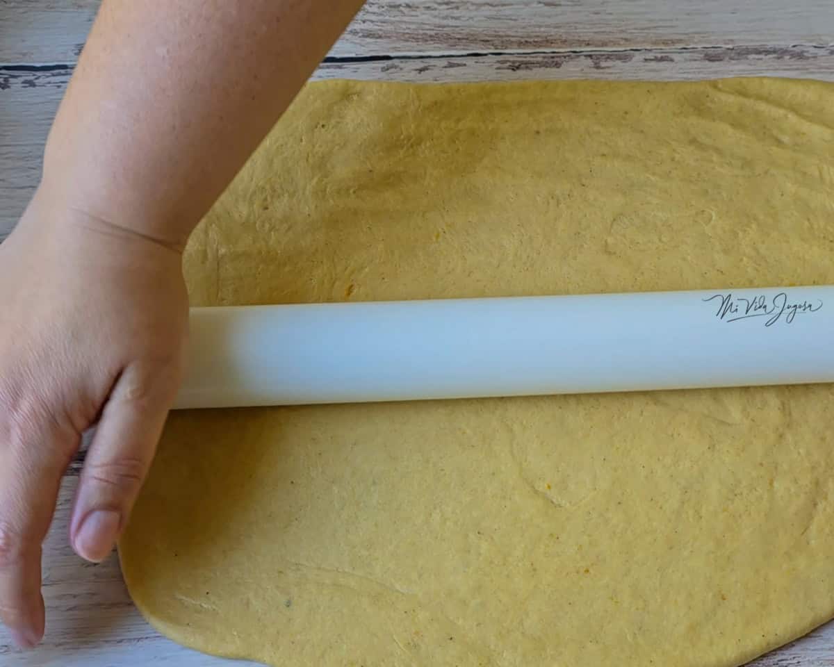 Una mano sostiene un rodillo mientras aplana una masa en forma rectangular sobre una superficie.