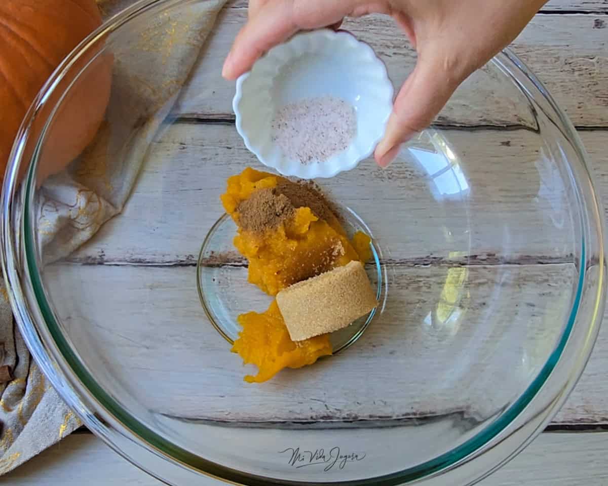 Un tazón de vidrio con puré de calabaza y azúcar morena, mientras una mano por encima espolvorea sal desde una tacita.