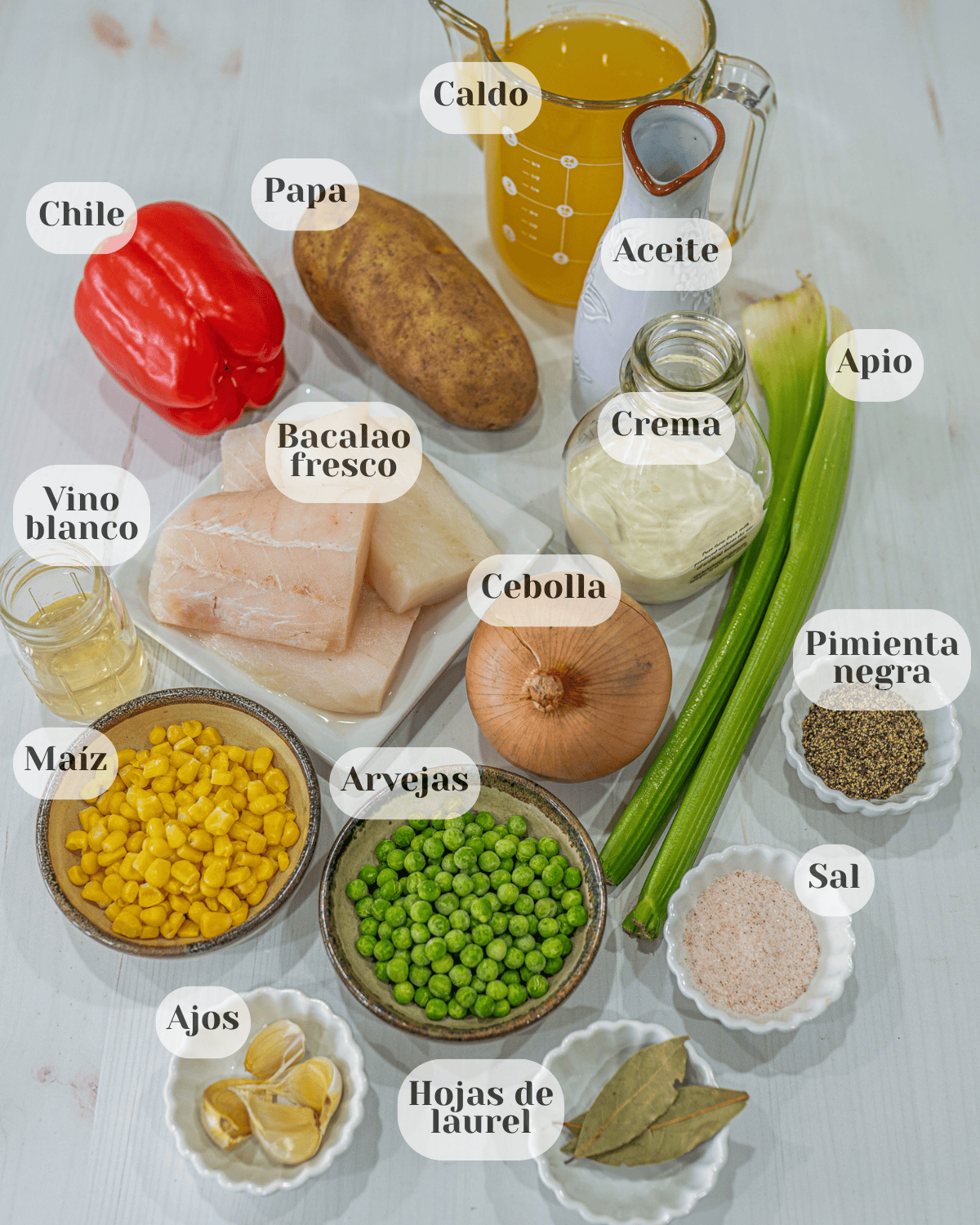 Una fotografia desde arriba donde se aprecian los ingredientes, una taza de caldo, una botellita de aceite, una botellita de crema dulce, dos tallos de apio, una papa, un chile morron, un platito con bacalao fresco, un recipiente con vino blanco, una cebolla, una tacita de pimienta negra, una tacita de sal, una taza de maix dulce, otra con arvejas, unos ajos y unas hojsa de laurel
