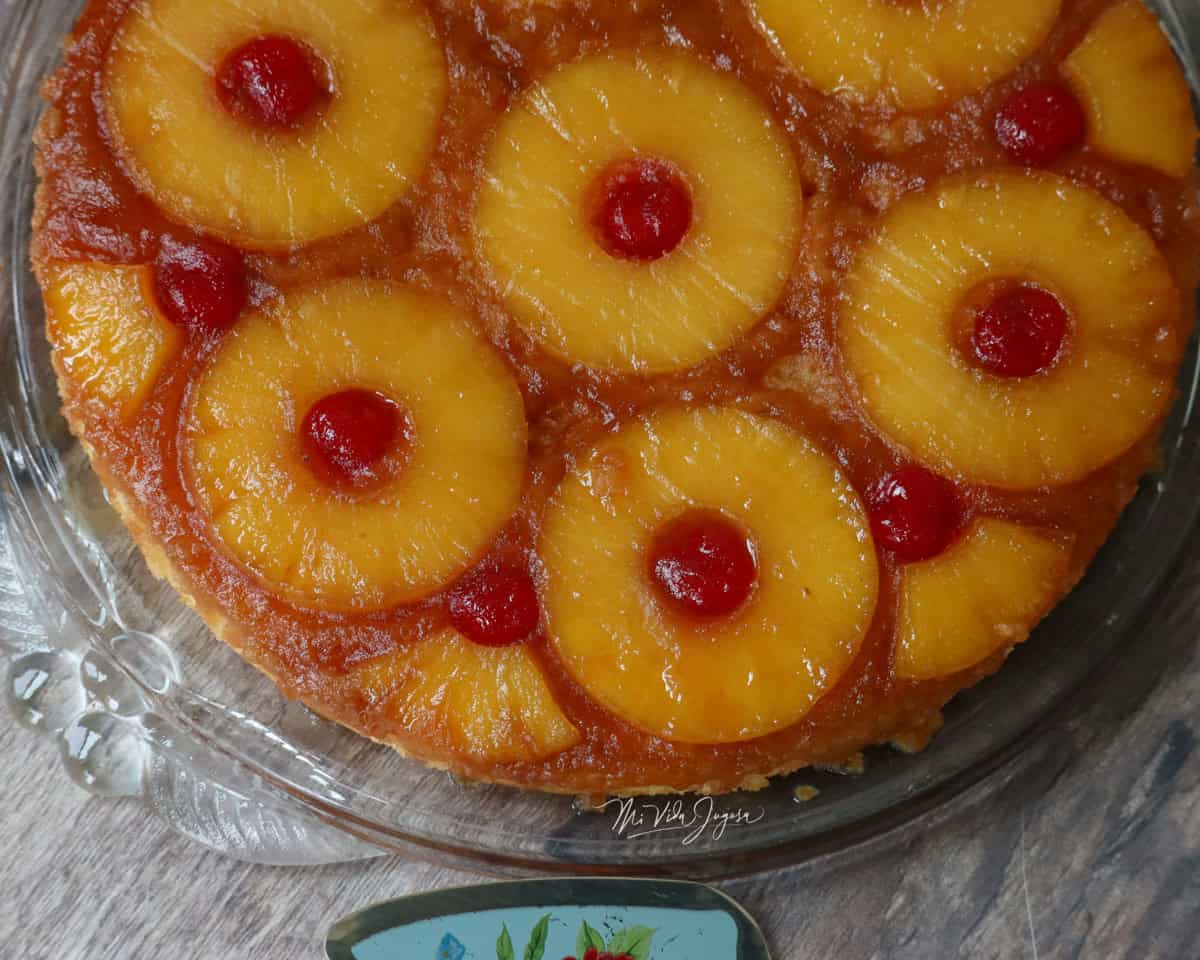 Un pastel redondo de volteado de piña, decorado por encima con rodajas de piña y cerezas, sobre un platon de vidrio.