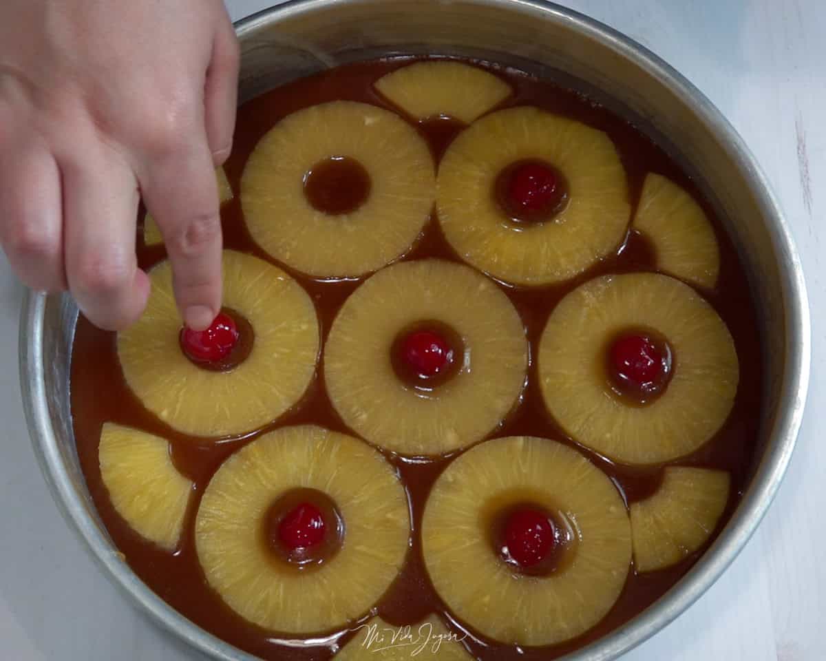 Un molde con una base de caramelo, mientras una mano acomoda pedazos de piña en forma de flor y coloca cerezas en el centro de cada rodaja.