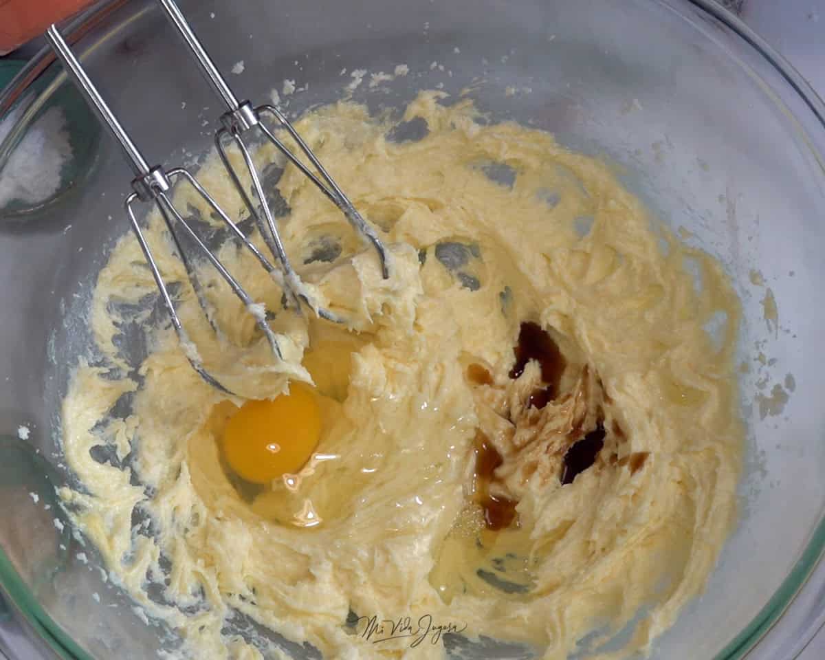 Un tazón con azúcar, mantequilla, dos huevos y esencia de vainilla, mientras se levantan ligeramente las aspas de la batidora con parte de la mezcla.