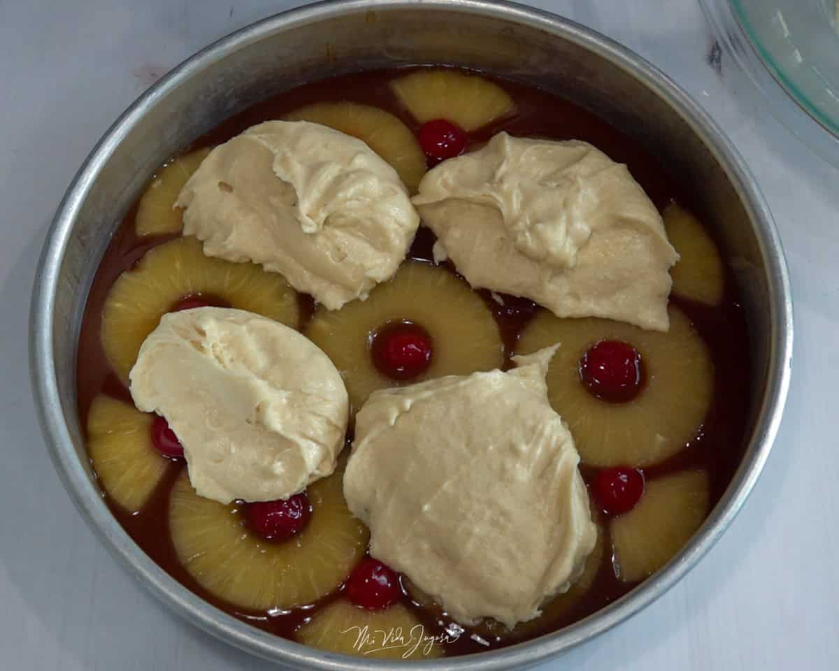 Un molde con el fondo cubierto de caramelo, decorado con piñas y cerezas, mientras por encima se colocan porciones de la mezcla del bizcocho.