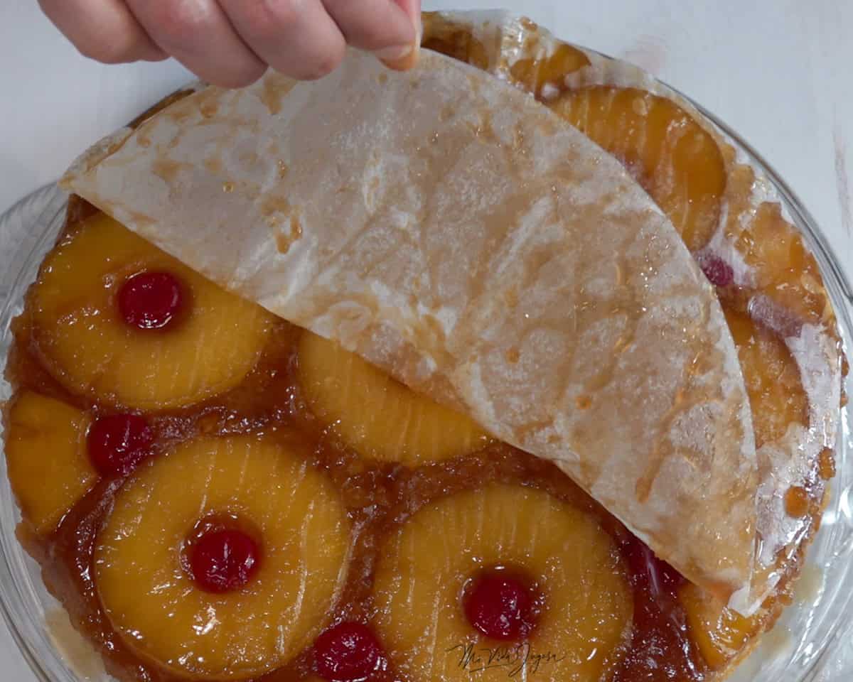 Un volteado de piña decorado por encima con rodajas de piña y cerezas, mientras una mano retira con cuidado el papel encerado de la superficie.