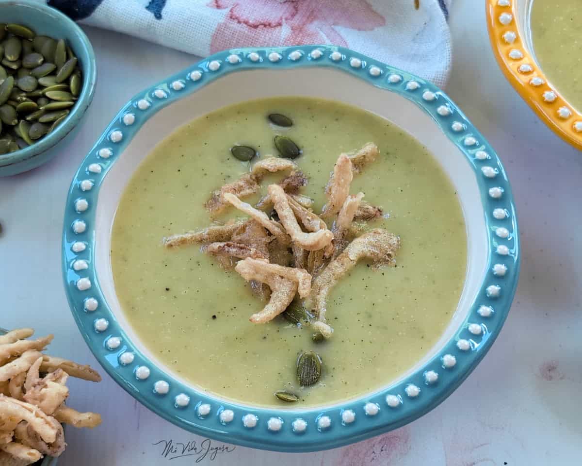 Una taza servida con crema de vegetales decorada con semillas de girasol y cebolla frita por encima. Al lado una taza conteniendo semillas de girasol, un taza de cebollas fritas y un limpion decorador.