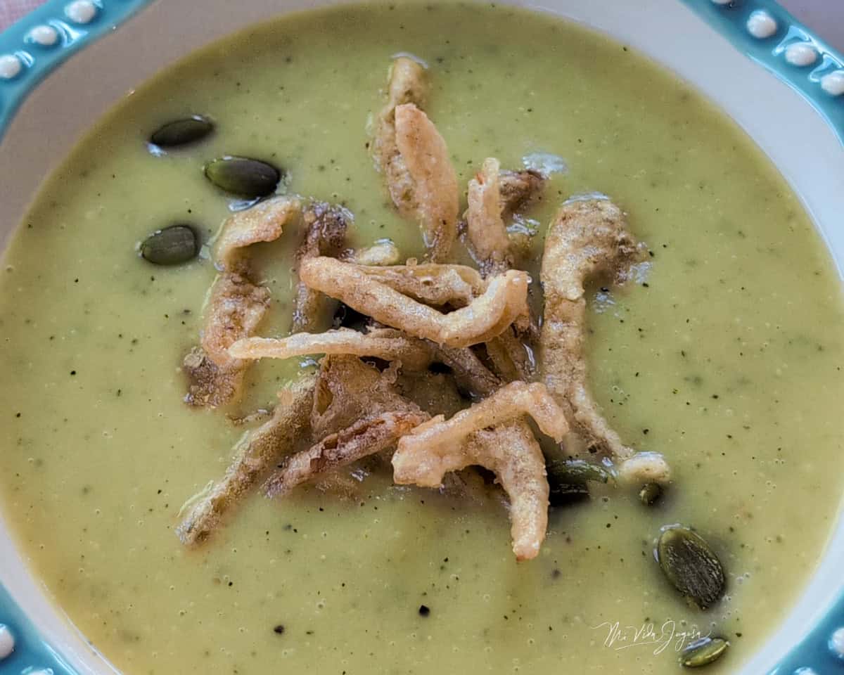 Un plato hondo con una cremosa crema de vegetales de color verde suave, decorada por encima con semillas de girasol y cebolla frita crujiente.