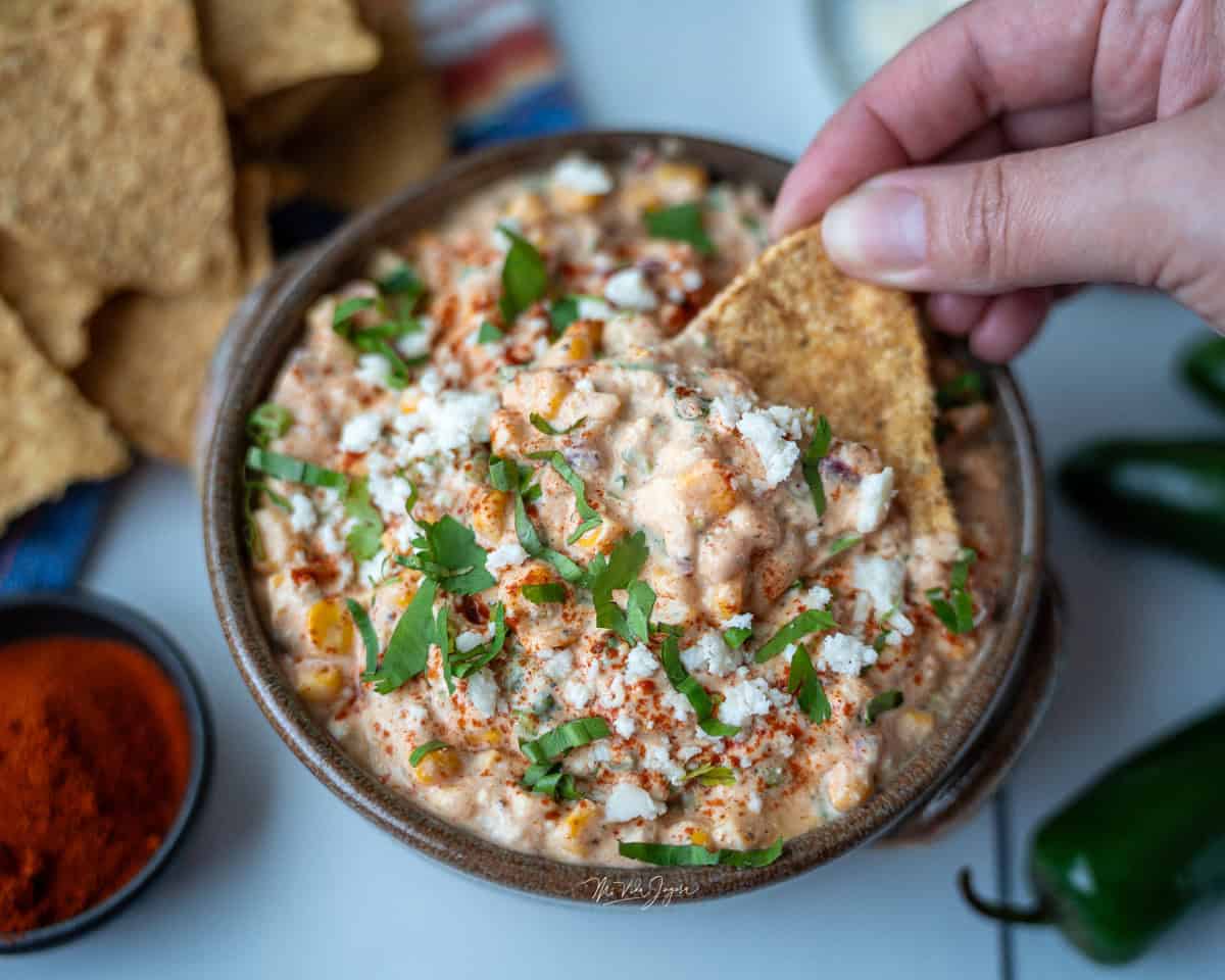 Una taza honda con dip de maíz, decorado con páprika por encima, culantro picado y queso. La foto está tomada desde arriba; a un lado se observan tortillas tostadas, una tacita pequeña con páprika y unos chiles jalapeños enteros, mientras una mano sostiene un chip con dip.
