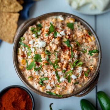 Una taza honda con dip de maíz, decorado con páprika por encima, culantro picado y queso. La foto está tomada desde arriba; a un lado se observan tortillas tostadas y una tacita pequeña con páprika.