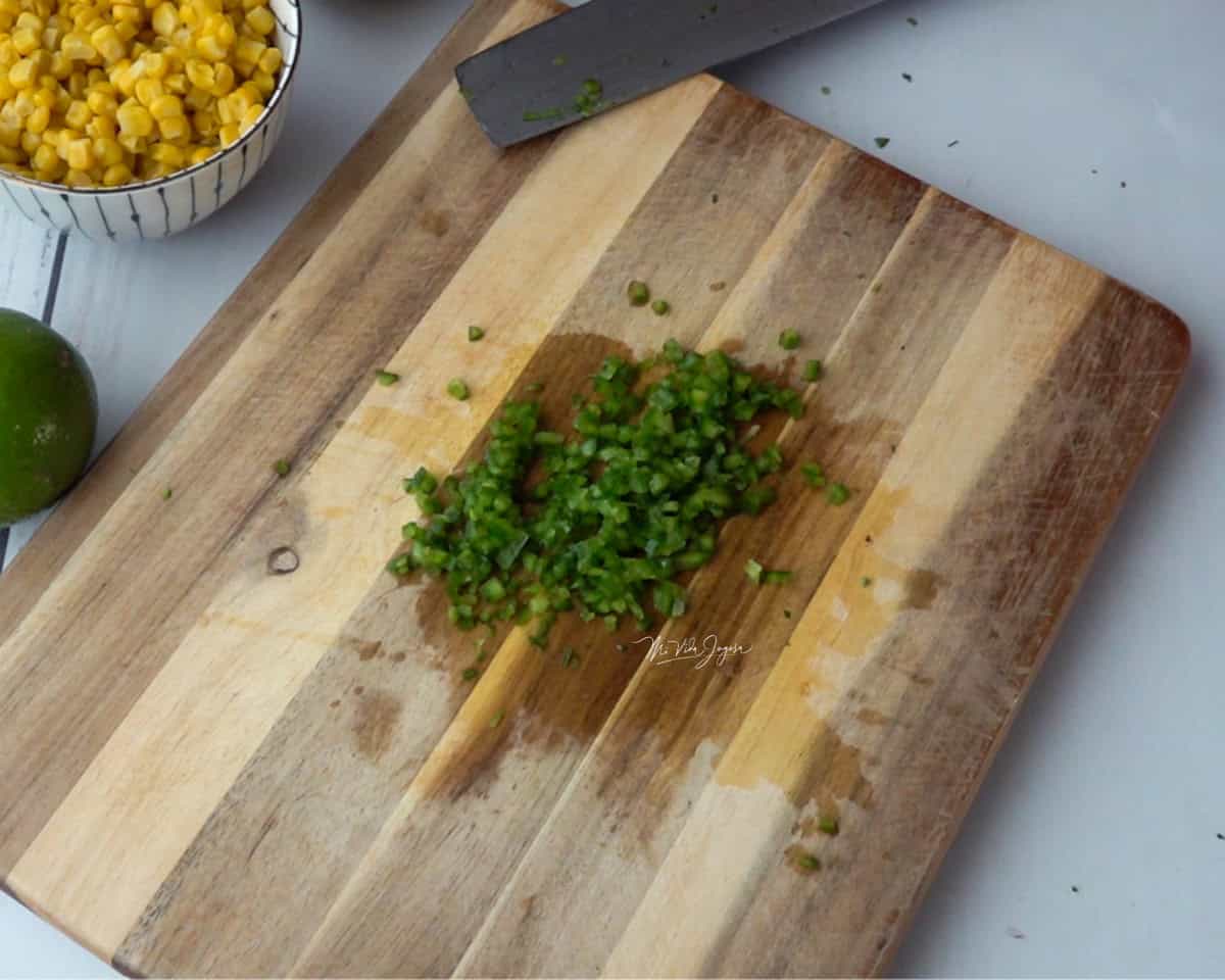 Una tabla de picar con un cuchillo al lado. Sobre la tabla se observa un jalapeño picado en cuadritos, y a un lado hay una taza con maíz dulce y un limón.