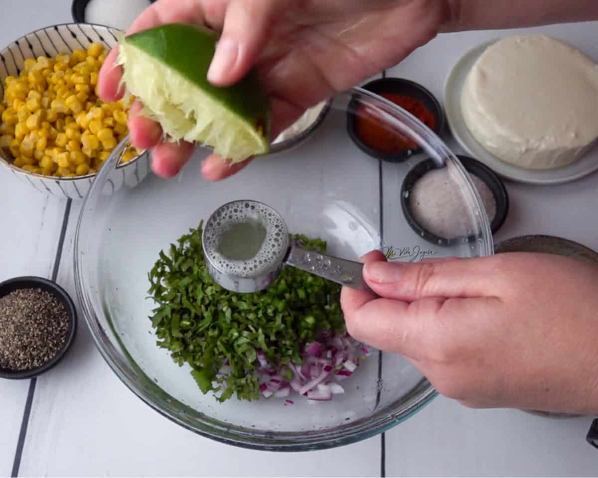 Un tazón con cebolla, culantro y cilantro. Encima se ven dos manos: una sostiene una taza de medida y la otra exprime medio limón para agregar el jugo dentro de la taza. Al lado del tazón se observan el maíz, el queso y los condimentos en tacitas.