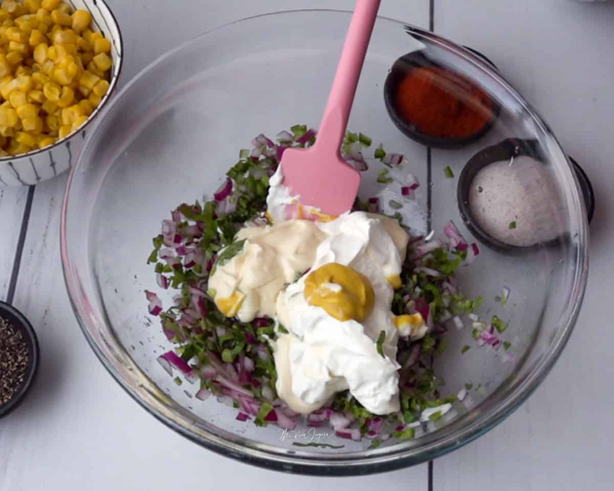 Un tazón con cebolla, culantro, cilantro, chile jalapeño, natilla, mayonesa y mostaza, con una espátula dentro. Al lado del tazón se observa maíz dulce en una taza, páprika y sal.