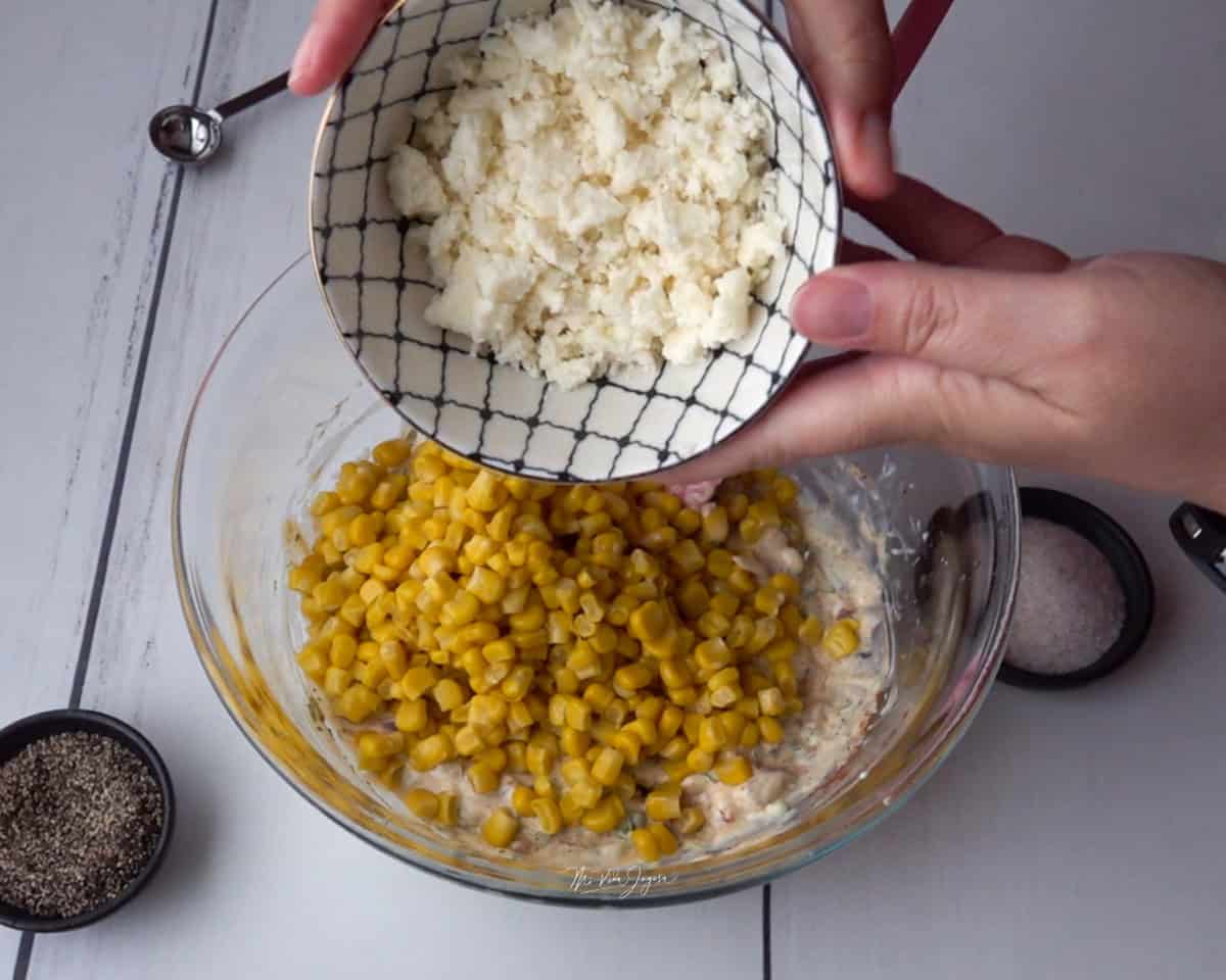 Un tazón con la mezcla del dip y maíz por encima. Sobre el tazón, dos manos sostienen una taza con queso fresco. A un lado se observan una tacita de sal y pimienta negra.