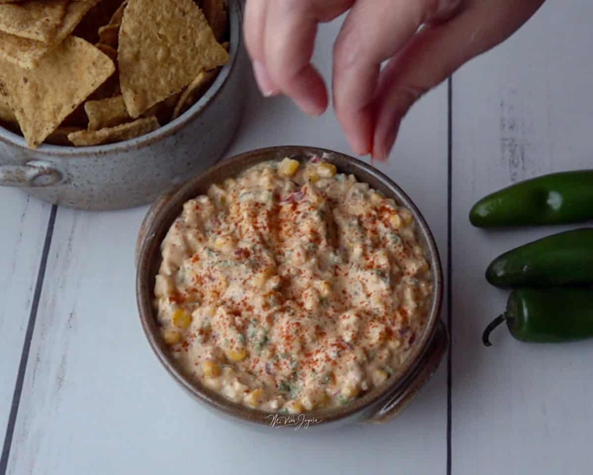Una taza con dip de maíz, unos chiles jalapeños al lado y una taza con chips. Una mano espolvorea páprika por encima del dip.