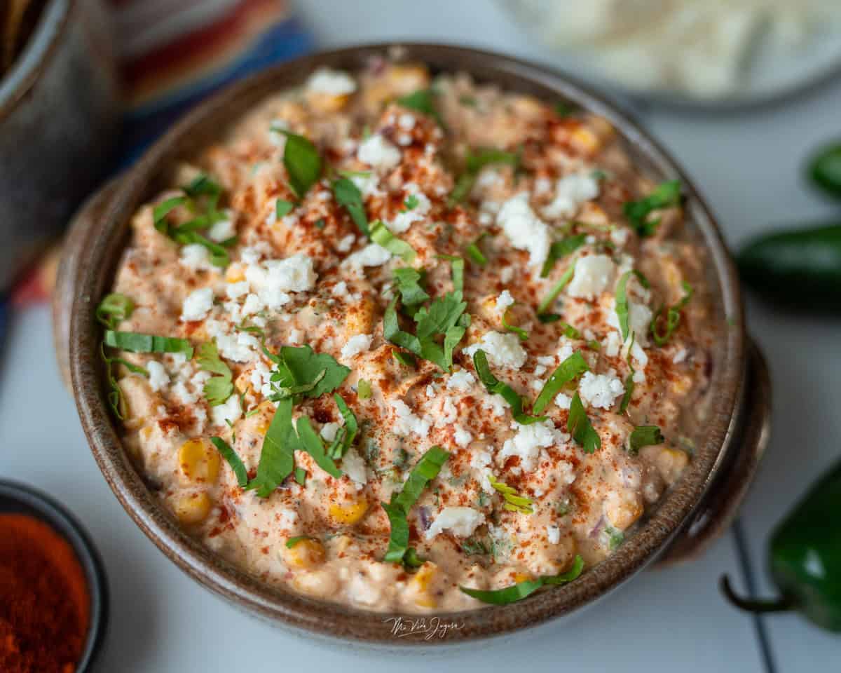 Una taza honda con dip de maíz, decorado con páprika por encima, culantro picado y queso. La foto está tomada desde arriba; a un lado se observan tortillas tostadas y una tacita pequeña con páprika.