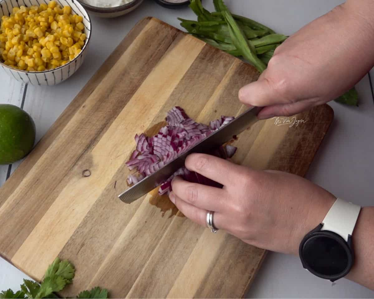 Una tabla de picar con una cebolla morada siendo cortada. Una mano sostiene la cebolla y la otra sostiene el cuchillo. Alrededor se ve una tacita con maíz dulce, un rollo de culantro y un limón.