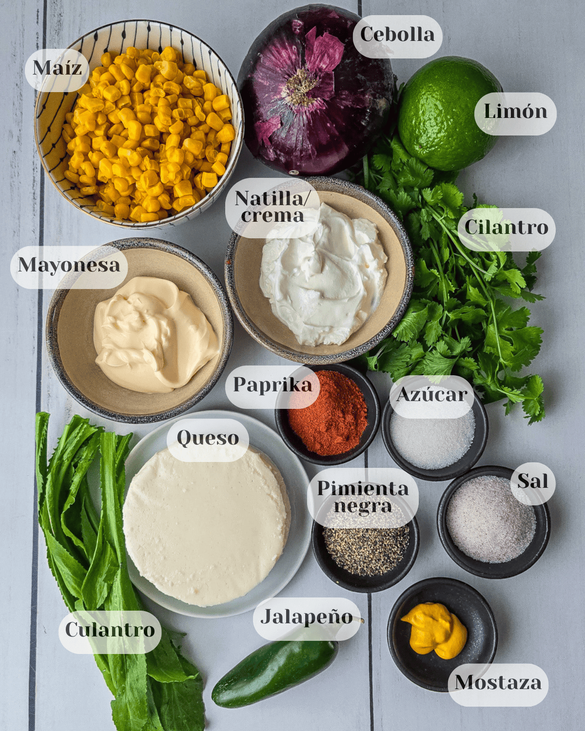 Foto tomada desde arriba con los ingredientes para preparar el dip: una taza de maíz dulce, una cebolla morada entera, un limón entero, un rollo de culantro y cilantro, una tacita de mayonesa, otra de natilla, páprika, azúcar, pimienta negra, sal, mostaza, un platito con queso fresco redondo y un chile jalapeño.