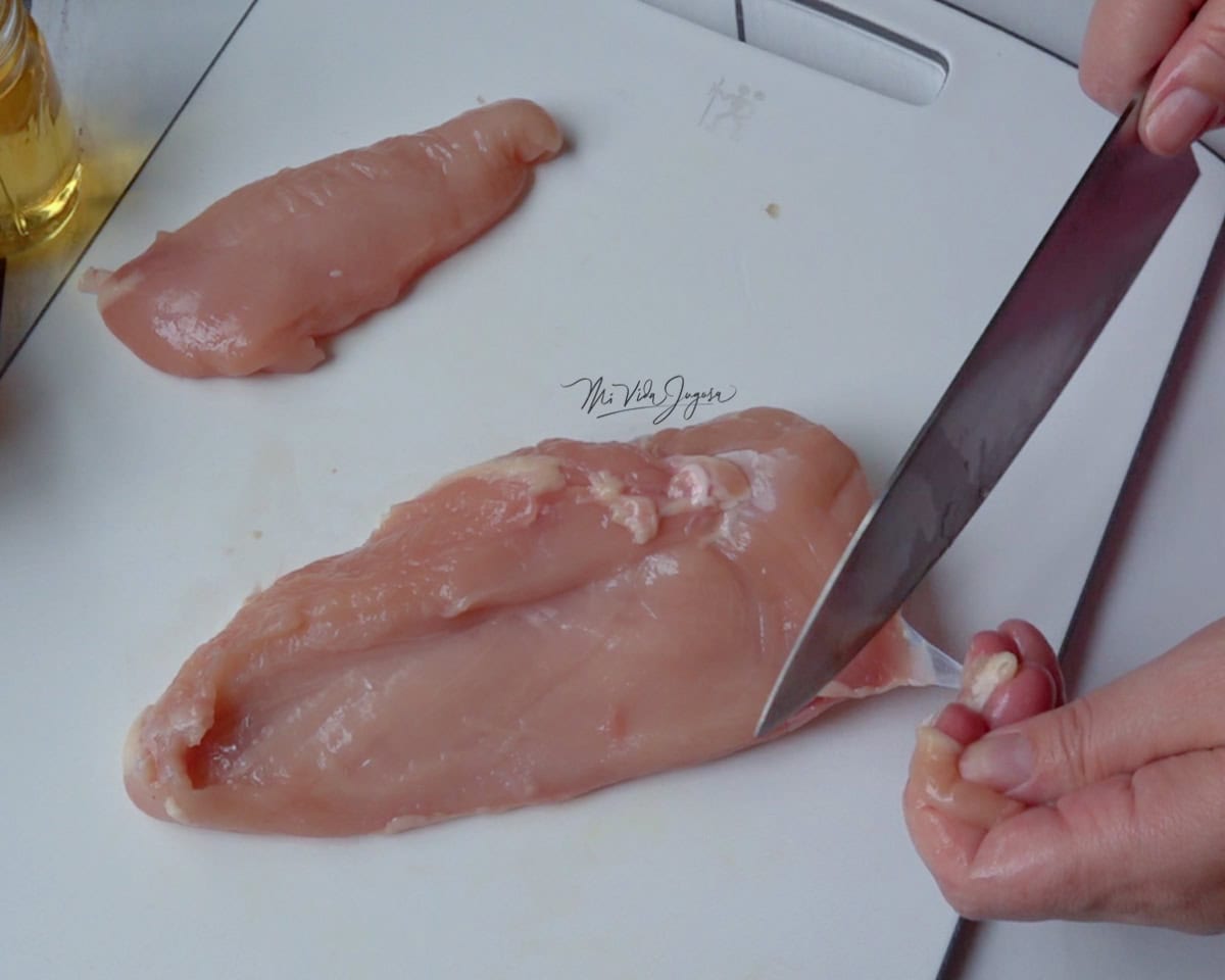 una tabla de picar blanca, encima una pechuga de pollo. La mano izquierda sostiene un pedazo de pollo, mientras la derecha corta los excesos de grasa en la pechuga