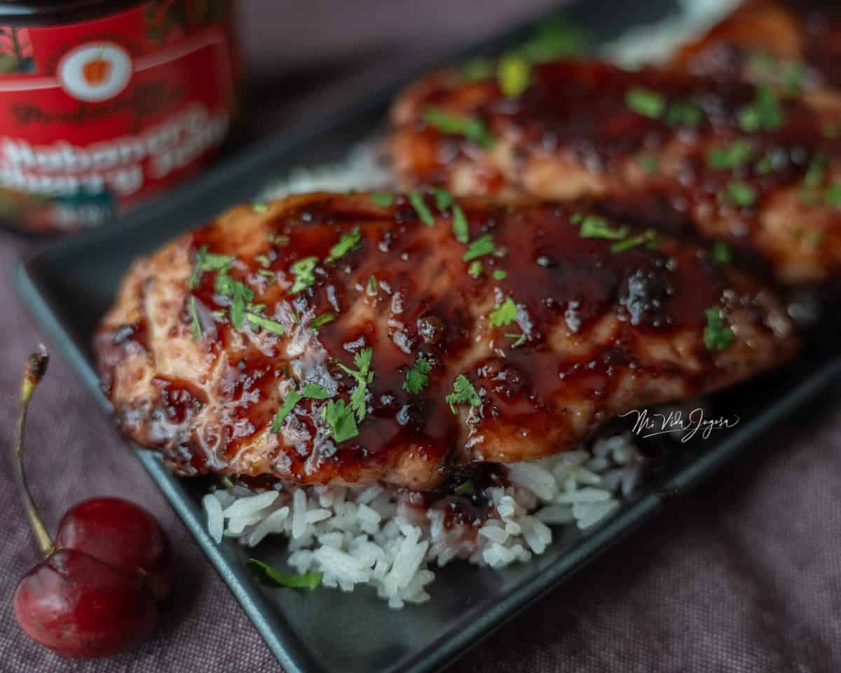 un plato cuadrado negro, con una porcion de arroz blanco con dos pedazos de pollo con mermelada de cereza, la mermelada tiene un color oscuro