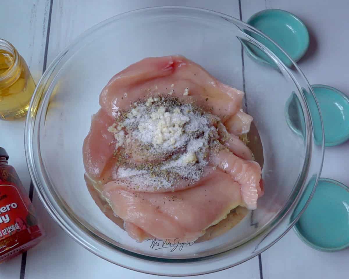 Un tazon de vidrio transparente, que contiene los pedazos pequeños de pollo crudo, encima se observa el aceite, y las especias utilizadas para marinar el pollo
