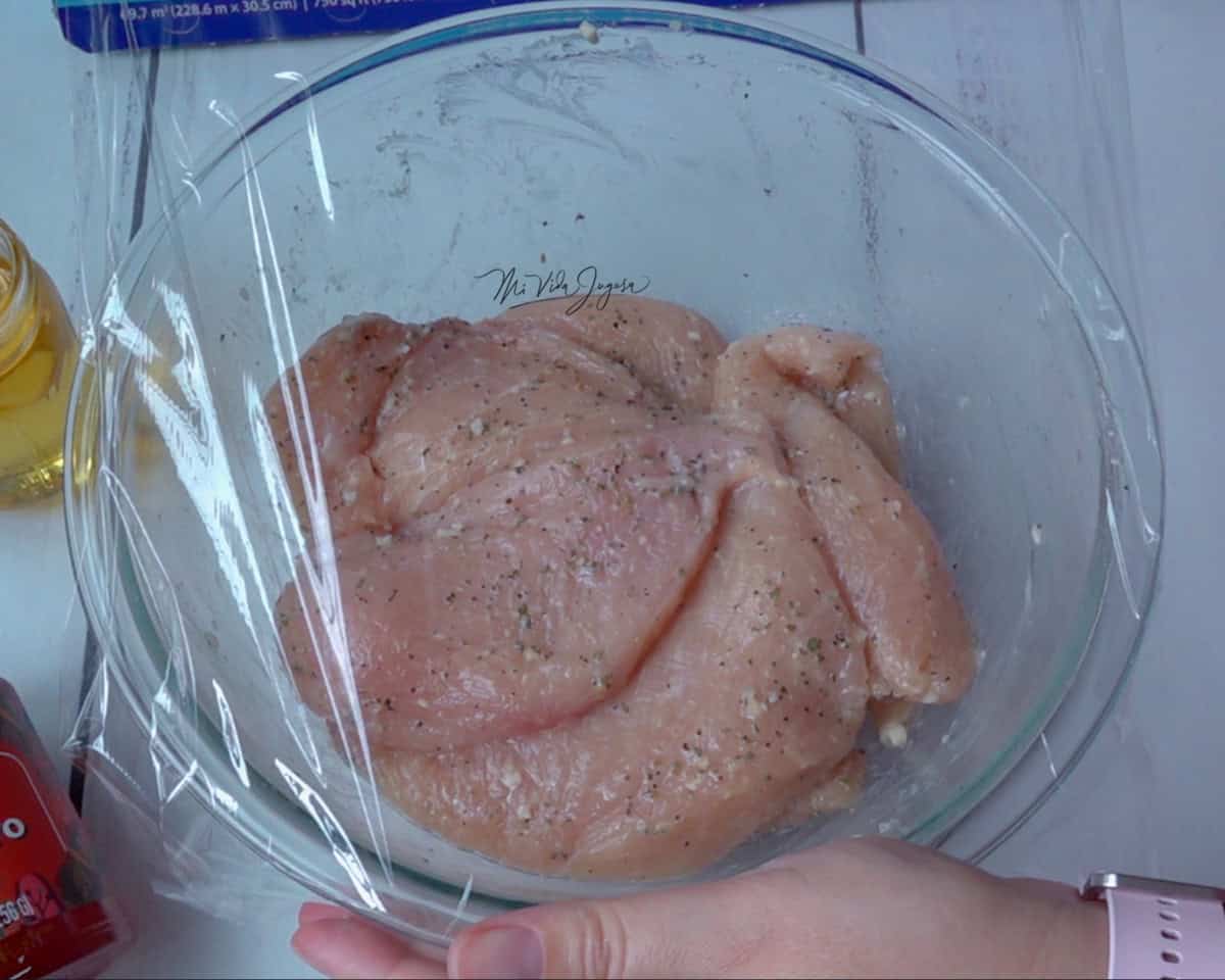 Un tazon de vidrio transparente que contiene el pollo marinado, se observa una capa delgada de plastico de cocina se coloca sobre el tazón, para cubrir el pollo y llevarlo a refrigeracion