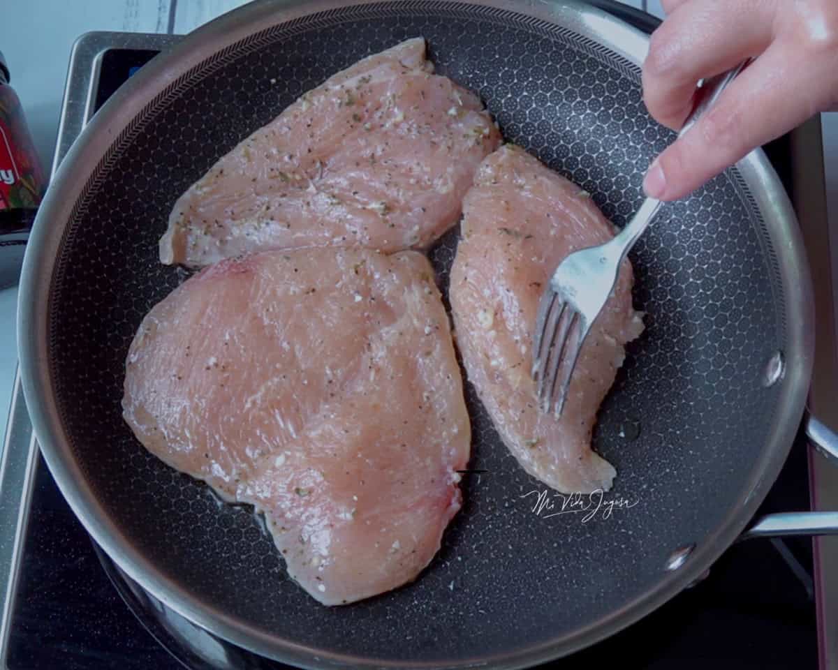 Un sarten de acero inoxidable que contiene 3 pedazos de pollo, se observa una mano derecha girando el polllo para cocinarlo por el otro lado