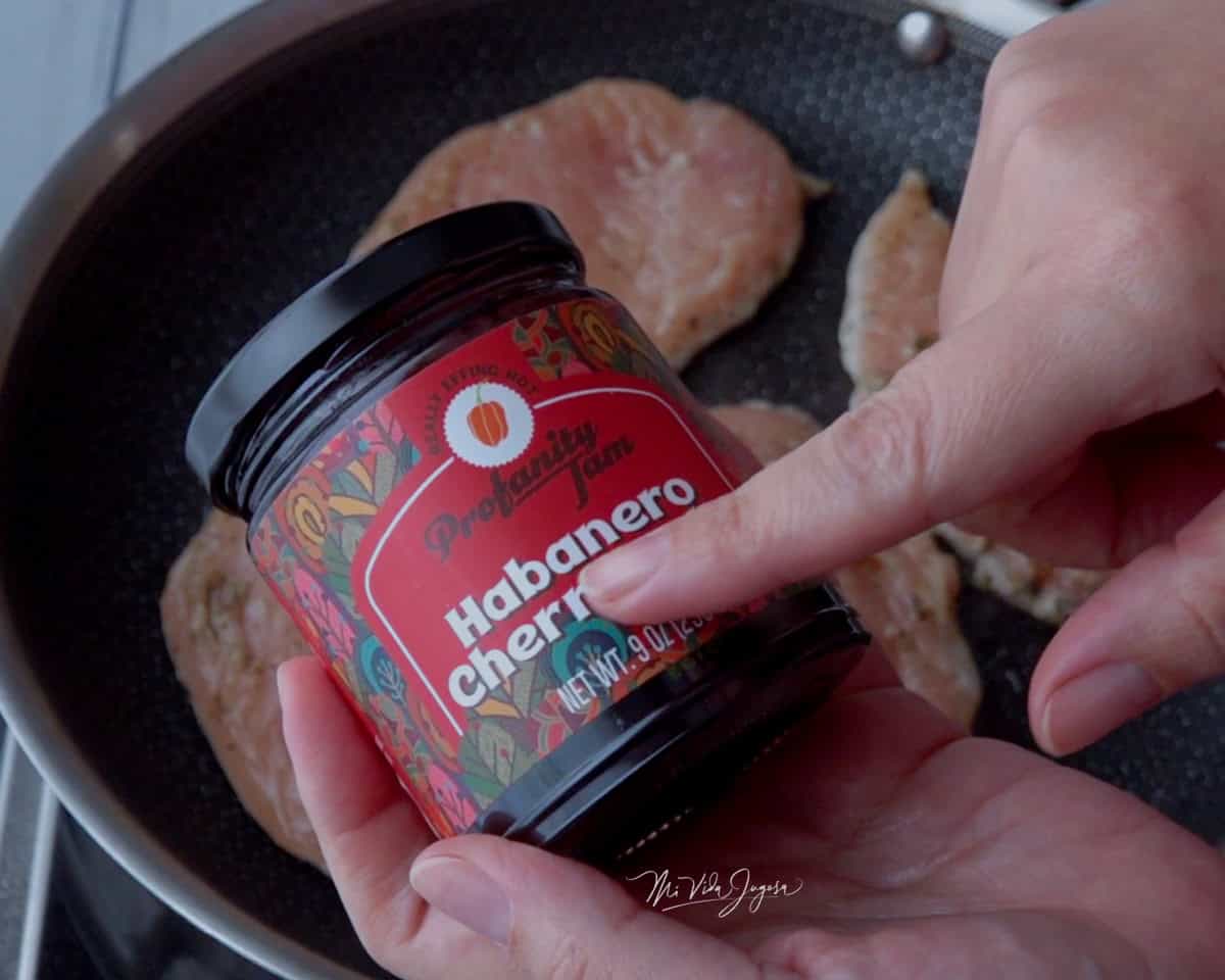 Se observa a lo lejos sarten de acero inoxidable que contiene 3 pedazos de pollo, mas cerca dos manos conteniendo un envase de mermelada