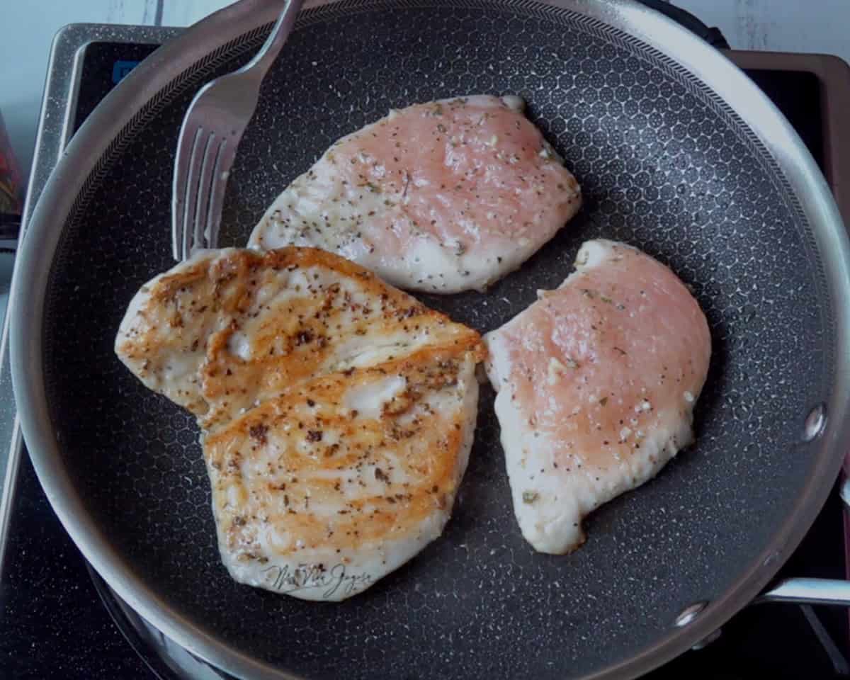 un sarten de acero inoxidable, donde se cocinan tres pedazos de pollo, 1 de los pedazos se ha girado y se observa el color dorado del asado, mientras los otros dos pedazos se observan crudos