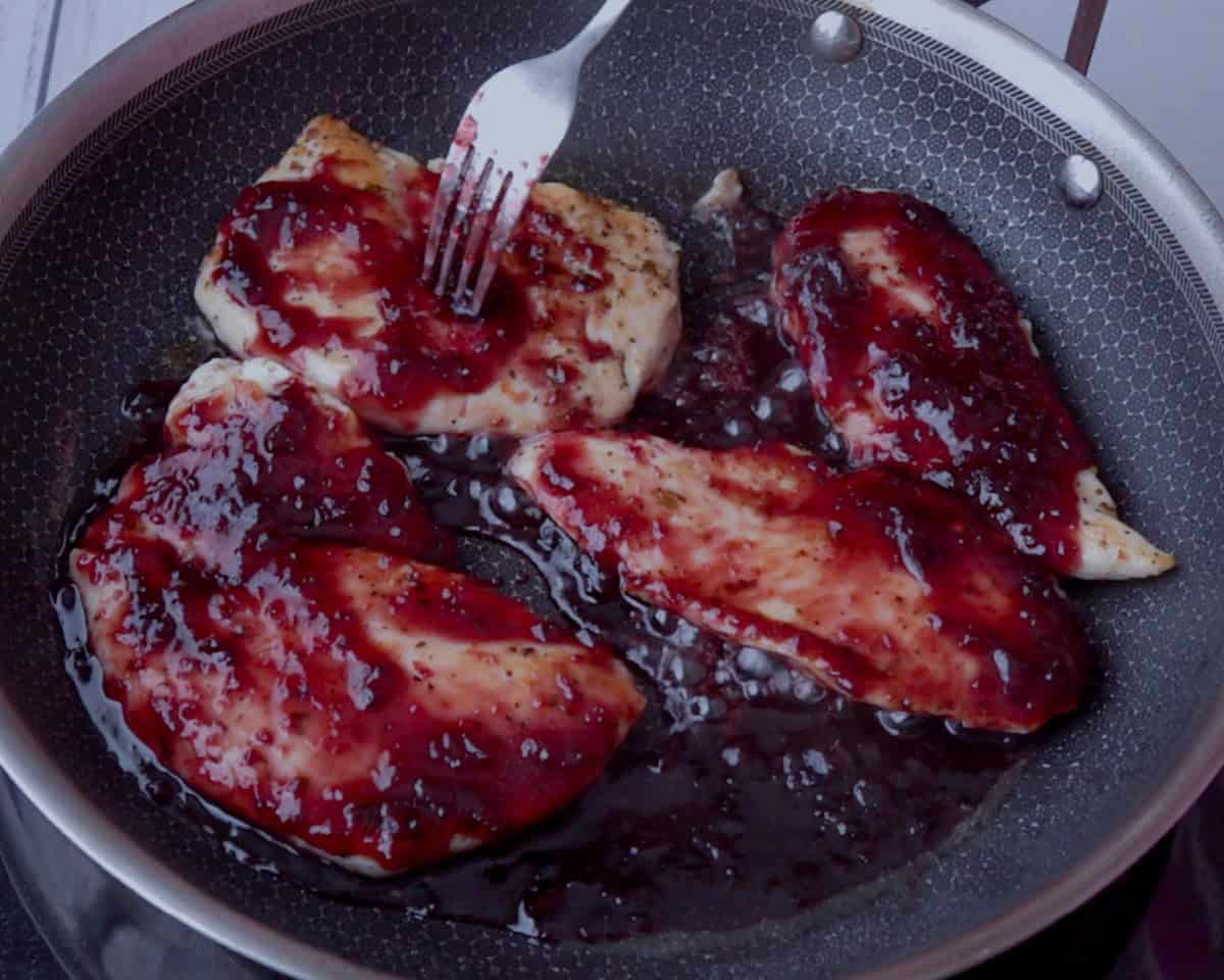 Cuatro pedazos de pollo sobre un sarten de acero inoxidable, se observa donde se estan cocinando con mermelada de cereza, con un color vivo de la mermelada