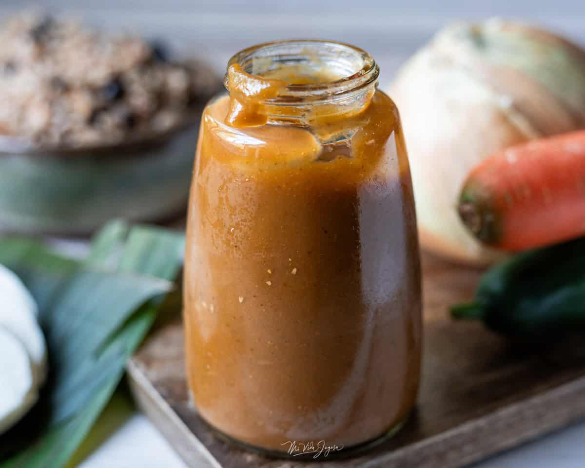 Una botella de vidrio con salsa Lizano casera sobre una tabla de madera, junto a cebolla, zanahoria y chile dulce, y al fondo un plato con gallo pinto.