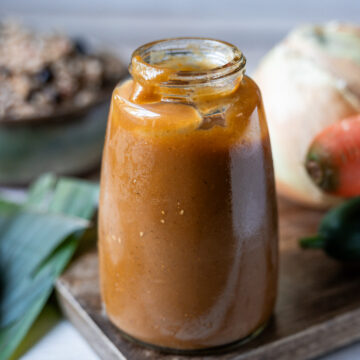 Una botella de vidrio con salsa Lizano casera sobre una tabla de madera, junto a cebolla, zanahoria y chile dulce, y al fondo un plato con gallo pinto.