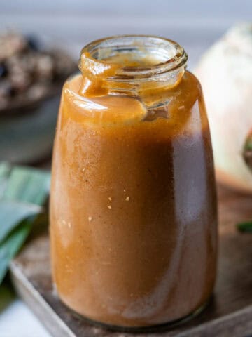 Una botella de vidrio con salsa Lizano casera sobre una tabla de madera, junto a cebolla, zanahoria y chile dulce, y al fondo un plato con gallo pinto.