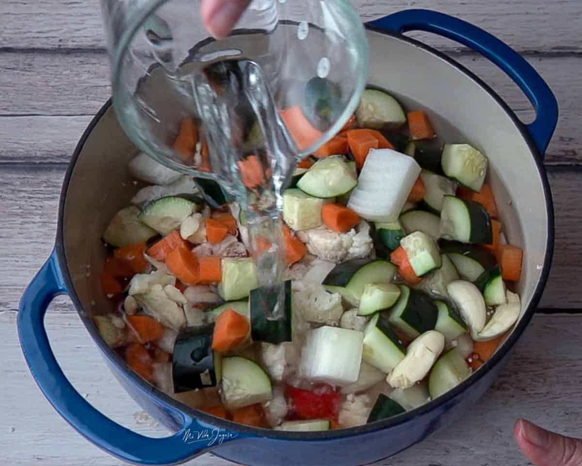 Una olla con vegetales picados en cuadritos mientras una taza agrega agua.