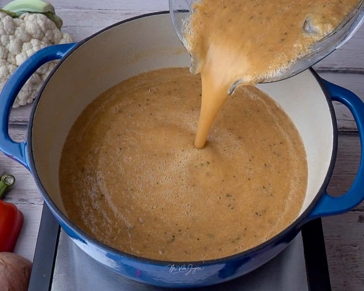 Una olla sobre una plantilla de cocina mientras el pichel de la licuadora vierte la mezcla de vegetales licuados.