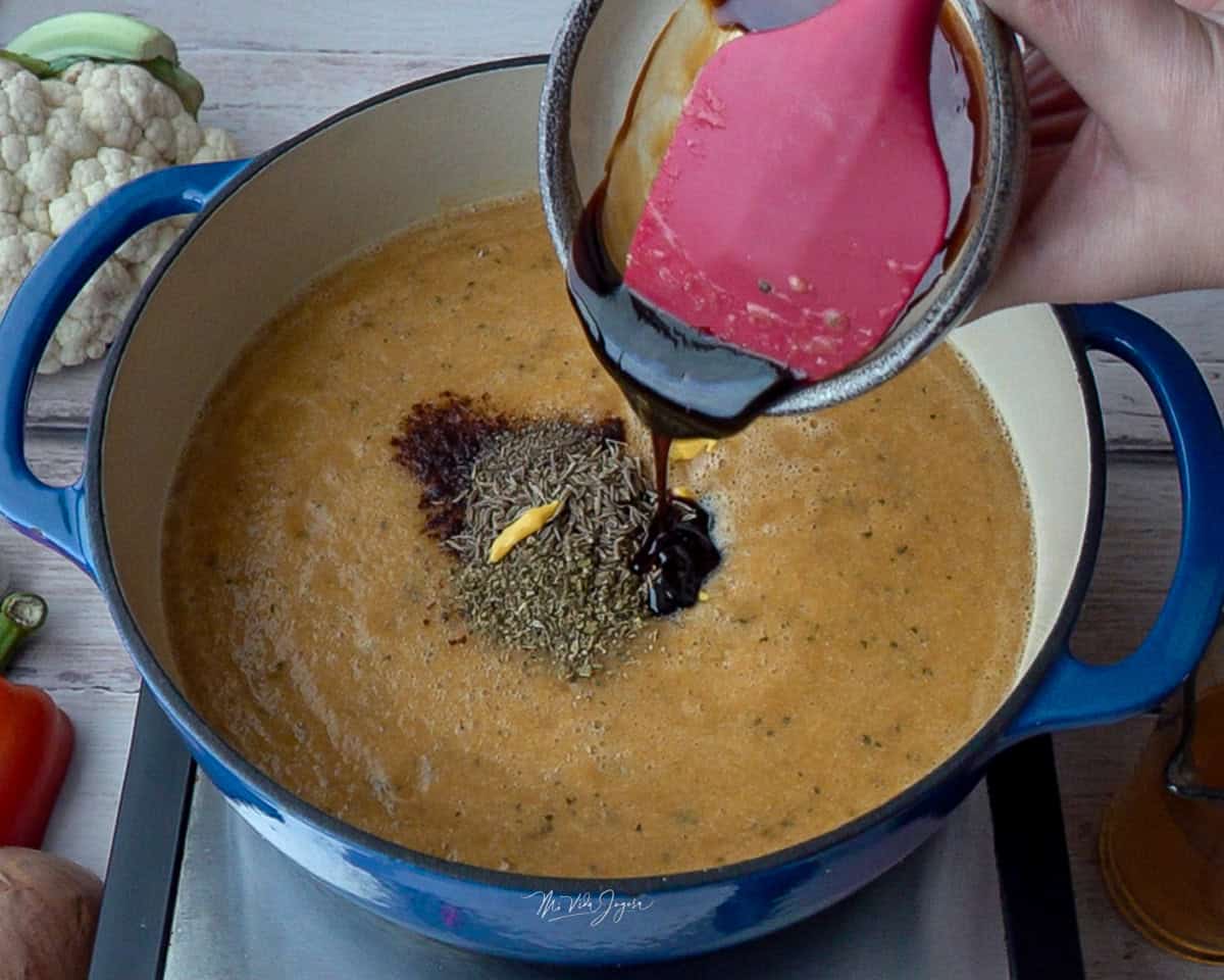 Una olla con la mezcla de vegetales licuados y los condimentos encima, mientras una espátula agrega la melaza desde una tacita.