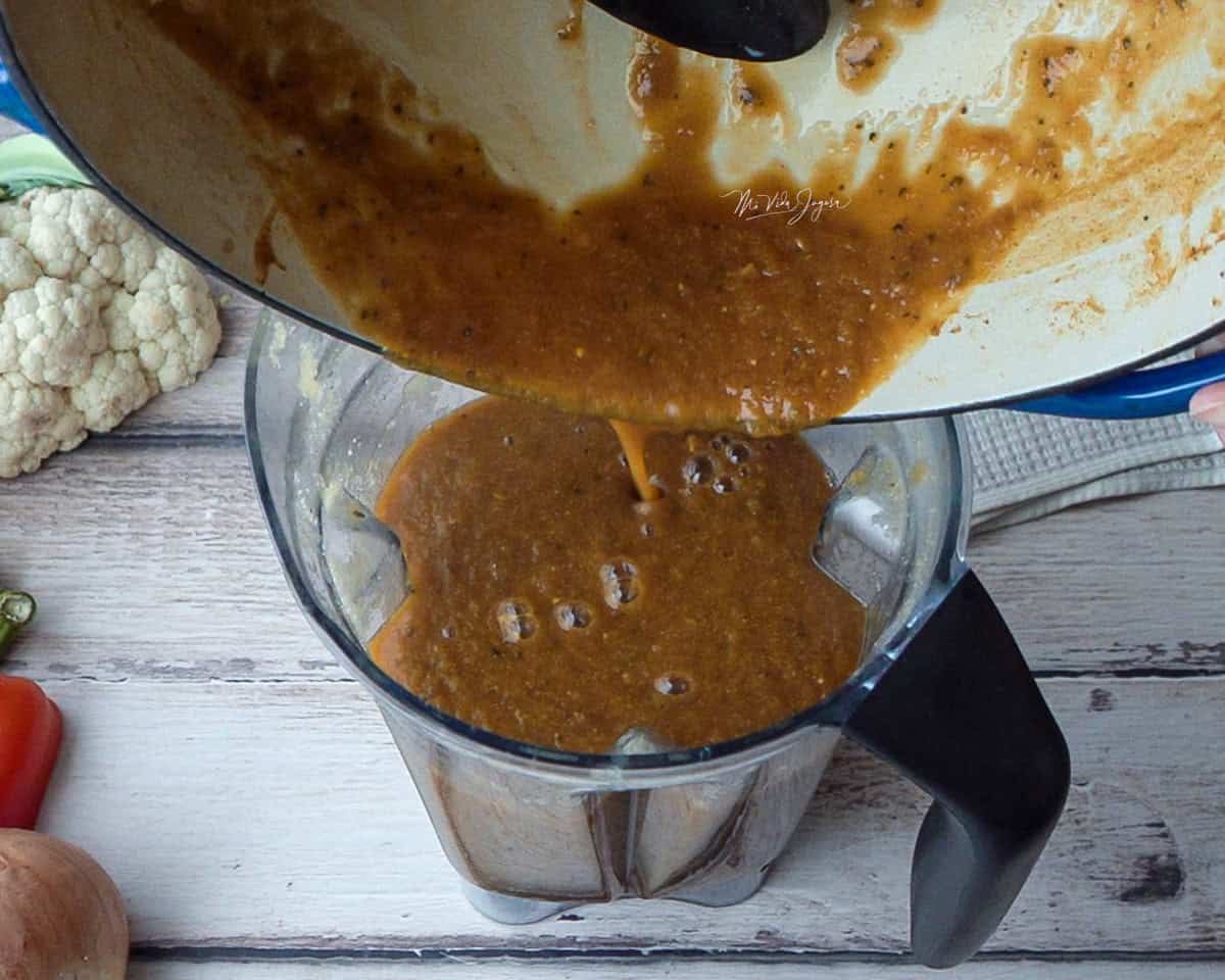 Una olla vierte la mezcla en el vaso de la licuadora; afuera se observan una coliflor, un chile dulce y una cebolla.