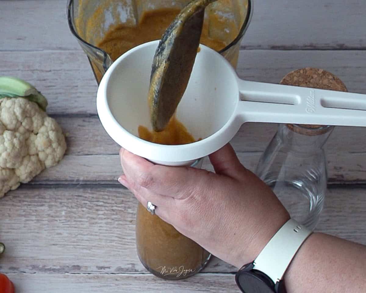 Una mano sostiene un envase con un embudo encima, mientras la otra mano vierte la salsa con una cuchara. Al fondo se ve el pichel de la licuadora, una coliflor y un envase vacío a la par.