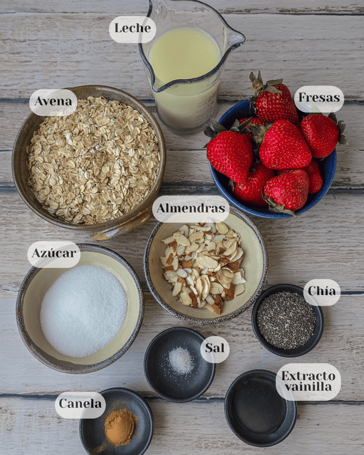 Una fotografía de vista aérea con todos los ingredientes organizados para preparar la avena trasnochada: leche en un tazón, avena en una taza, fresas en otra taza, almendras fileteadas, azúcar, chía, una pizca de sal, extracto de vainilla y canela.