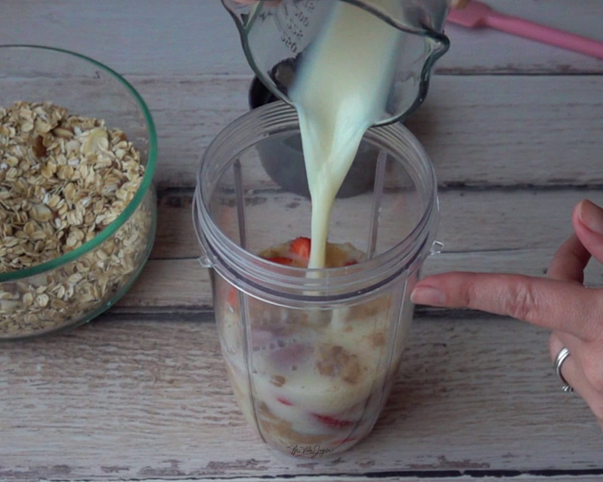 Un vaso de licuadora con fresas, azúcar, sal y extracto de vainilla, mientras se agrega leche desde un pichel en forma de chorro. Al lado se observa una taza con avena y, al fondo, una espátula.