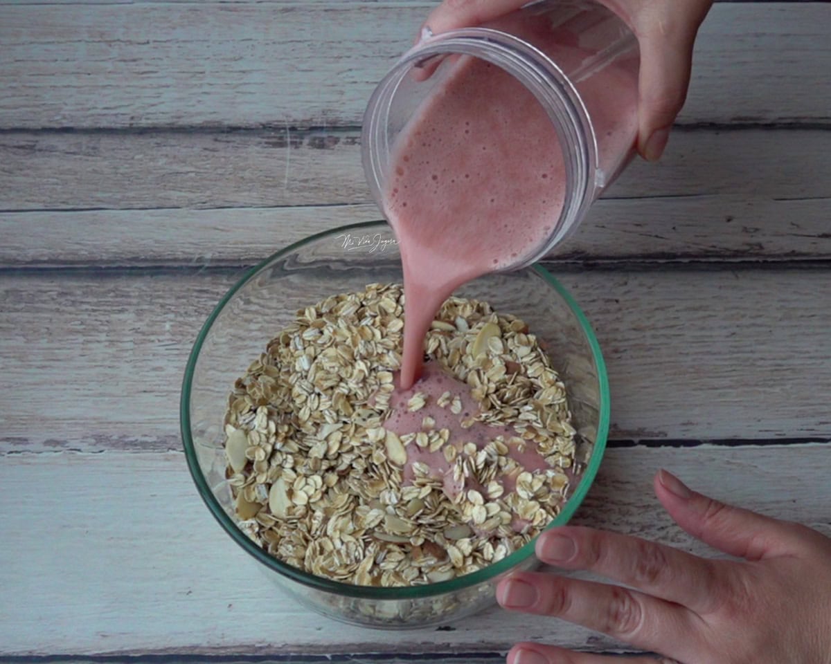 Una mano sostiene una taza con hojuelas de avena, mientras la otra sostiene el vaso de la licuadora con un batido de fresa y lo incorpora a la avena.