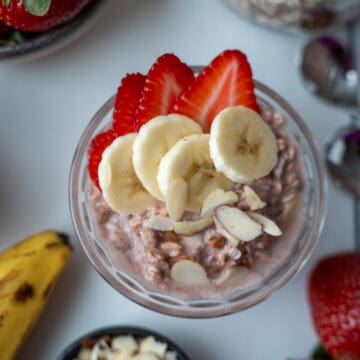 Una taza de vidrio con avena trasnochada, decorada con pedacitos de almendra, fresas y rodajas de banano. Al lado se observa un banano, varias fresas frescas y un poco de avena sin remojar.