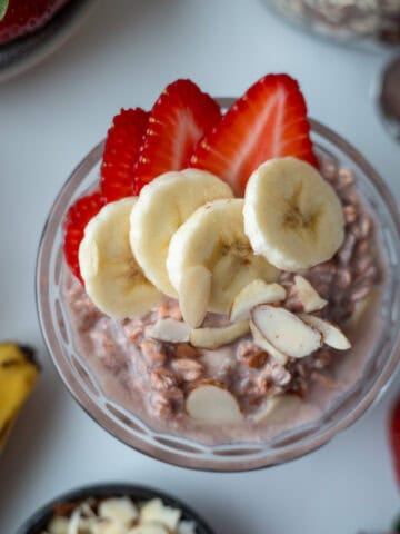 Una taza de vidrio con avena trasnochada, decorada con pedacitos de almendra, fresas y rodajas de banano. Al lado se observa un banano, varias fresas frescas y un poco de avena sin remojar.